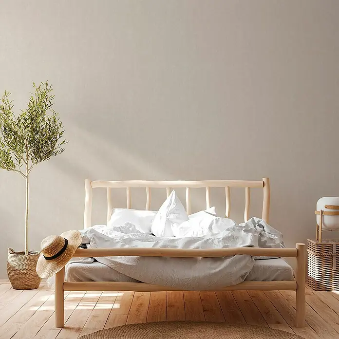 A.S. Création Casual Living Vliestapete - Harmonisches Beige, Uni, 10,05 x 0,53 m - Hellholz Bettgestell mit weißer Bettwäsche in einem hellen Raum mit Holzboden. Eine Pflanze und ein Hut stehen neben dem Bett.