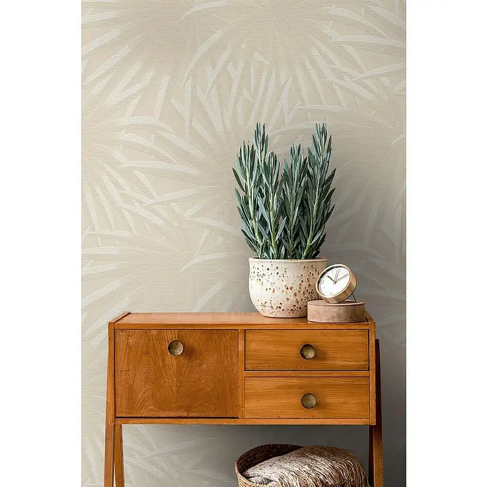 A.S. Création Casual Living Vliestapete Palmenblätter - Beige/Weiß, Floral, 10,05 x 0,53 m - Eine Nahaufnahme eines Sideboards mit einer Topfpflanze und einer dekorativen Uhr darauf, vor einer dezent gemusterten Wand.