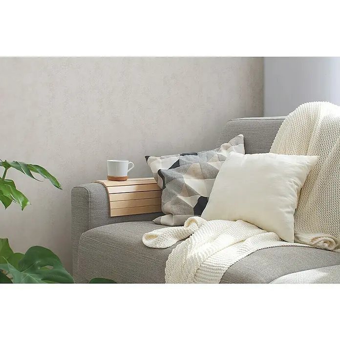 A.S. Création Casual Living Vliestapete - Creme, Uni, 10,05 x 0,53 m - Gemütliche Wohnzimmerszene mit einem grauen Sofa, dekoriert mit Kissen und einer Strickdecke, neben einer Monstera-Pflanze und einer Tasse.