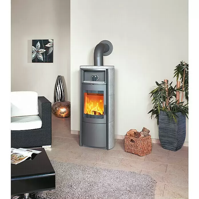 Hark Kaminofen Nika ECOplus - 7 kW, Raumheizvermögen: 193 m³, Grau - Moderner grauer Kaminofen mit sichtbaren Flammen, Steinplatte und Ablage, in einem Wohnzimmer mit Korb voller Holzscheite.