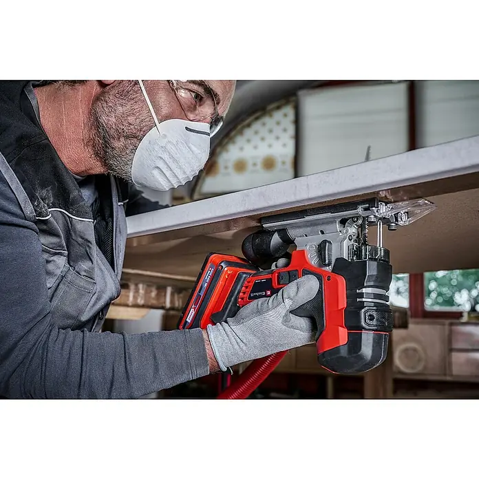 Einhell Professional Power X-Change 18V Akku-Pendelhubstichsäge TP-JST 18/135 Li BL - Solo - 18 V, Li-Ionen, Ohne Akku - Nahaufnahme einer Einhell Akku-Stichsäge in Aktion, bedient von einem Handwerker mit Schutzmaske. Das Gerät schneidet präzise in ein Holzbrett.