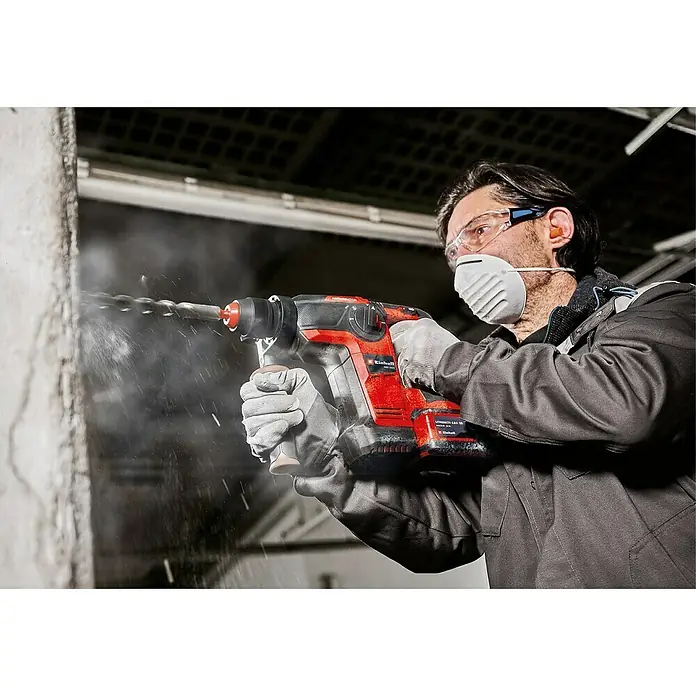 Einhell Professional Power X-Change 18V Akku-Kombihammer TP-HD 18/26 Li BL - Solo - 18 V, Ohne Akku, 0 Ah, 2,6 J, 1.200 U/min, Max. Schlagzahl: 5.500 /min - Ein Mann bohrt mit einer roten Einhell-Bohrmaschine in eine Wand, Schutzbrille und Maske tragend. Staub fliegt umher.