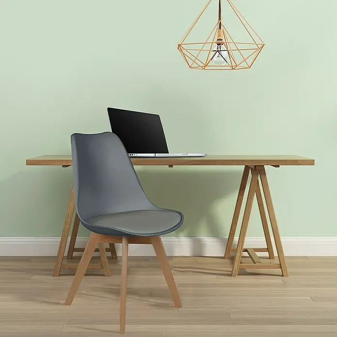 Home Deco Factory Silla Copenhague - Gris, 4 pzs. - Una silla gris con patas de madera delante de un escritorio de madera con un portátil y lámpara geométrica.
