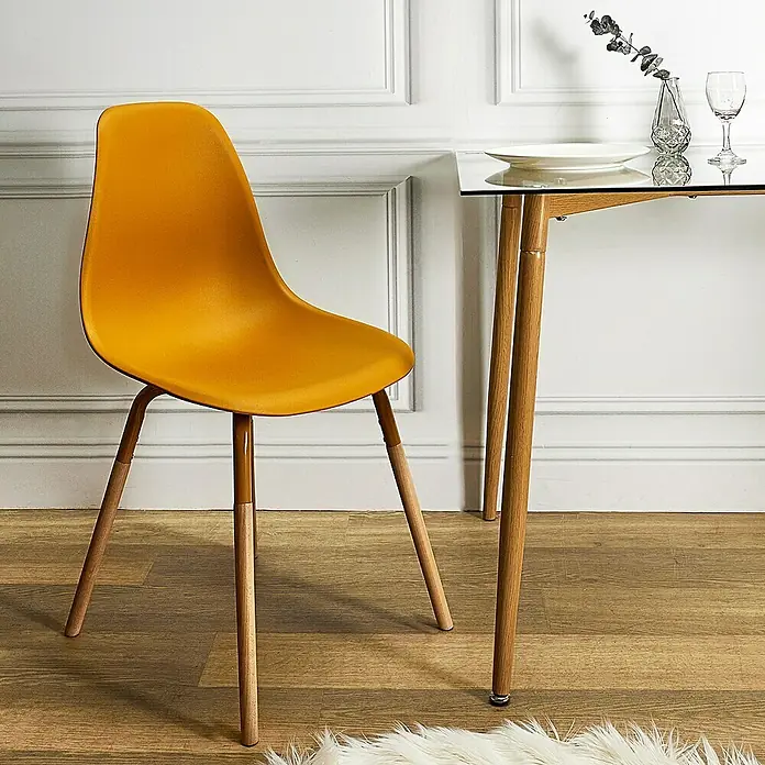 Silla Phenix - Mostaza, 4 pzs. - Silla amarilla con patas de madera al lado de una mesa con tapa de cristal contra una pared blanca.