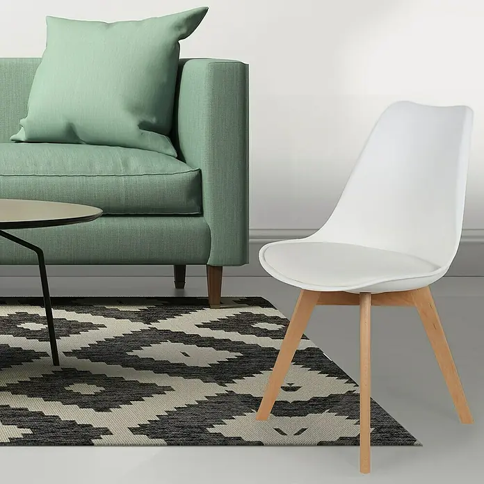 Silla Copenhague - Blanco, 4 pzs. - Carcasa blanca sobre una silla con patas de madera, junto a un sofá verde sobre una alfombra geométrica.
