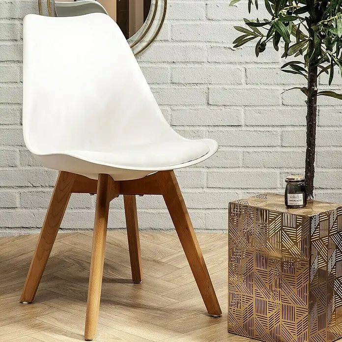 Silla Copenhague - Blanco, 4 pzs. - Una elegante silla blanca con patas de madera se encuentra junto a una mesa auxiliar dorada con una vela aromática.