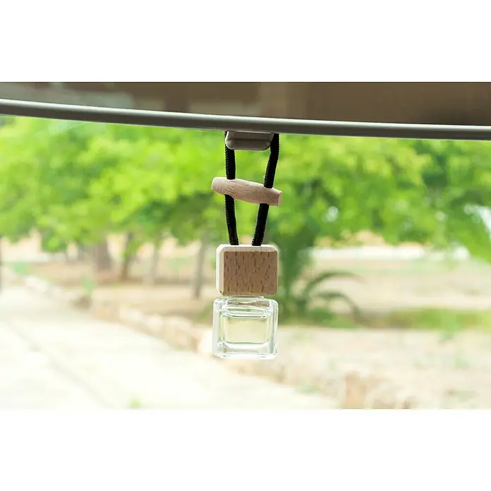 Ambientador de coche para colgar - Naranja, 8 sem. - Un difusor cuadrado de madera y vidrio con una cuerda negra cuelga delante de un fondo verde.