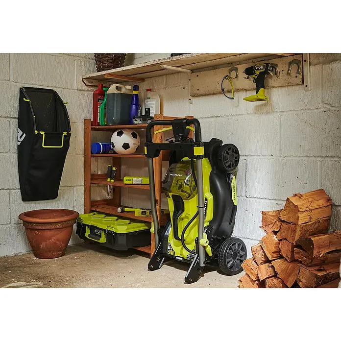 Ryobi ONE+ Akku-Rasenmäher RY18LMX40C-240 - 18 V, 2 Akkus, Schnittbreite: 40 cm - Aufnahme eines RYOBI Rasenmähers, der in einer Garage aufrecht gelagert wird, mit Werkzeugen und Gartenzubehör im Hintergrund.