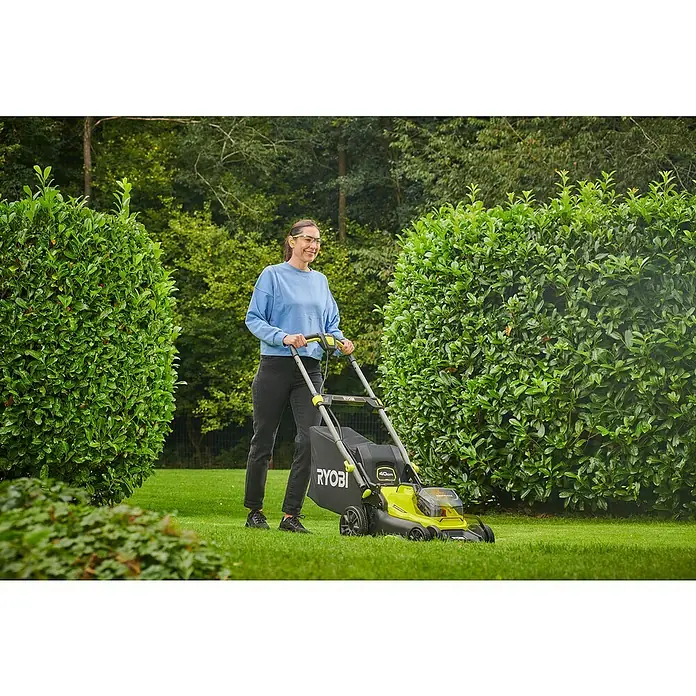 Ryobi ONE+ Akku-Rasenmäher RY18LMX40C-240 - 18 V, 2 Akkus, Schnittbreite: 40 cm - Frau benutzt einen RYOBI Akku-Rasenmäher auf einem Rasen zwischen zwei Büschen. Das Gerät ist gelb und schwarz mit einem Grasfangsack.