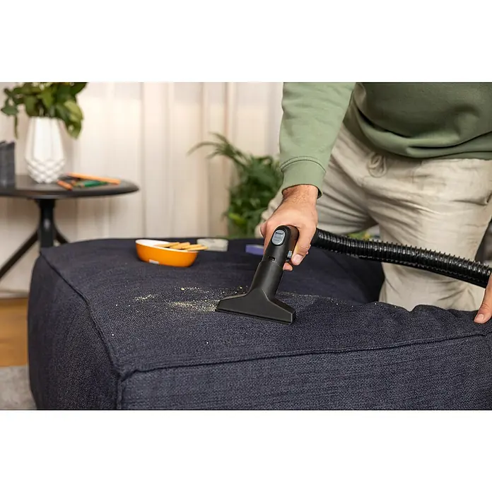 Kärcher Waschsauger SE 4 - 1.000 W, Behältervolumen: 4 l - Ein Mann saugt Krümel von einem blauen Polsterhocker mit einem schwarzen Handstaubsauger. Reinigungsgerät im Fokus.