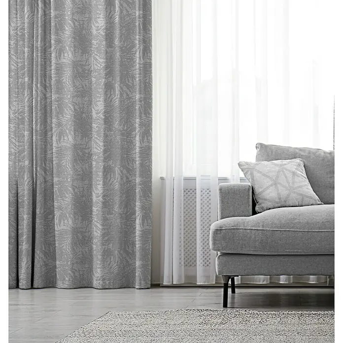 Cortina con ollaos Triana - An x Al: 140 x 270 cm, Gris - Una cortina gris con estampado de hojas junto a una ventana con cortinas translúcidas y un sofá gris, creando un ambiente acogedor.