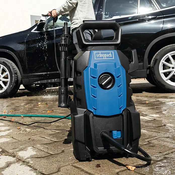 Scheppach Hidrolimpiadora de alta presión HPC1400 - 110 bar, 1.400 W, Capacidad de bombeo: 390 l/h - La imagen muestra una hidrolimpiadora Scheppach, utilizada en exteriores para tareas de limpieza. El dispositivo es azul y negro y se encuentra sobre una superficie pavimentada.