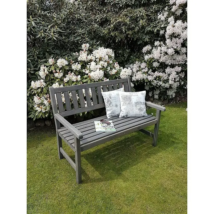 Promadino Gartenbank Johanna - B x T x H: 125 x 71 x 88,5 cm, Kiefer, Grau - Eine graue Gartenbank mit Dekokissen und Zeitschriften darauf, umgeben von blühenden Büschen auf einer grünen Wiese.
