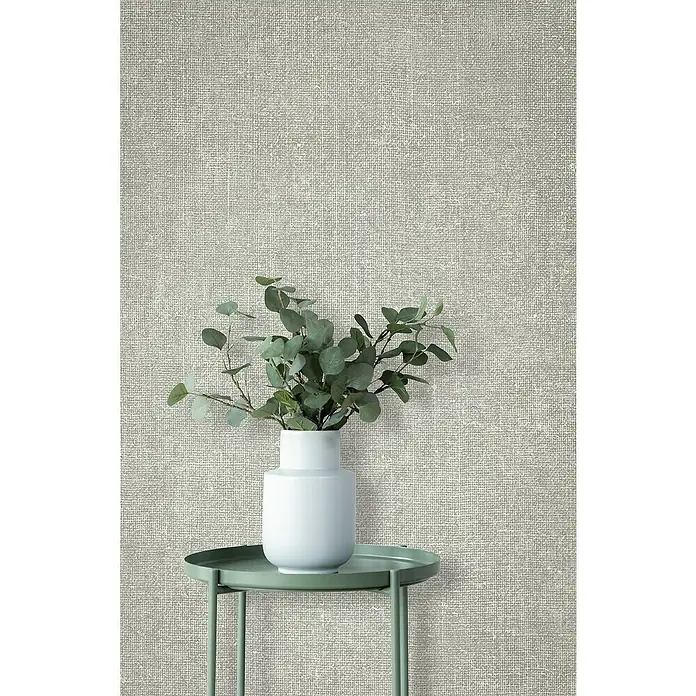 Marburg Struktura Vliestapete Struktur - Grün/Grau, Uni, 10,05 x 0,53 m - Nahaufnahme einer Tapete mit Leinenstruktur, die eine warme und natürliche Atmosphäre schafft. Ein weißer Vase mit Eukalyptuszweigen steht auf einem grünen Tisch.