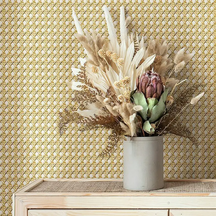 Marburg Struktura Vliestapete Holzgeflecht - Beige, Holzoptik, 10,05 x 0,53 m - Dekorative Trockenblumen in einer grauen Vase auf einem Holzschrank vor einem Hintergrund mit Rattanmuster.