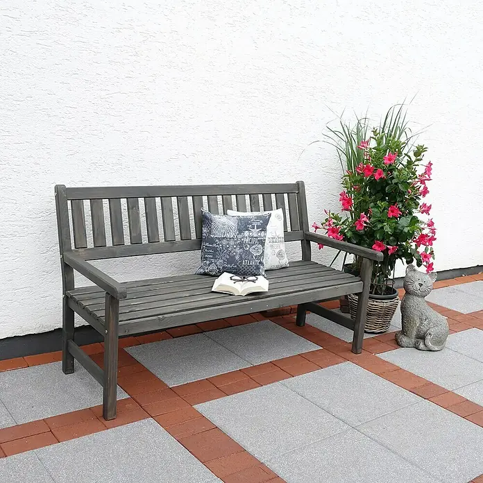 Promadino Gartenbank Johanna - B x T x H: 154 x 71 x 88,5 cm, Kiefer, Grau - Eine graue Holzbank mit Kissen und einer Pflanze im Topf vor einer weißen Wand. Die Bank steht auf einem gepflasterten Boden.