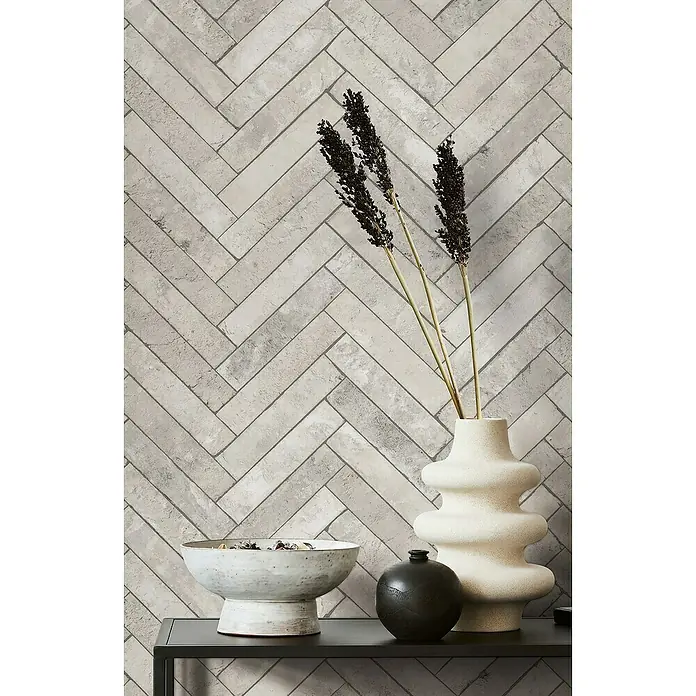Marburg Struktura Vliestapete Fischgrat - Grau, Steinoptik, 10,05 x 0,53 m - Nahaufnahme des stilvollen Interieurs mit grauem Ziegelmuster-Hintergrund, einer Vase mit Dekozweigen und Keramikschalen auf einem dunklen Tisch.