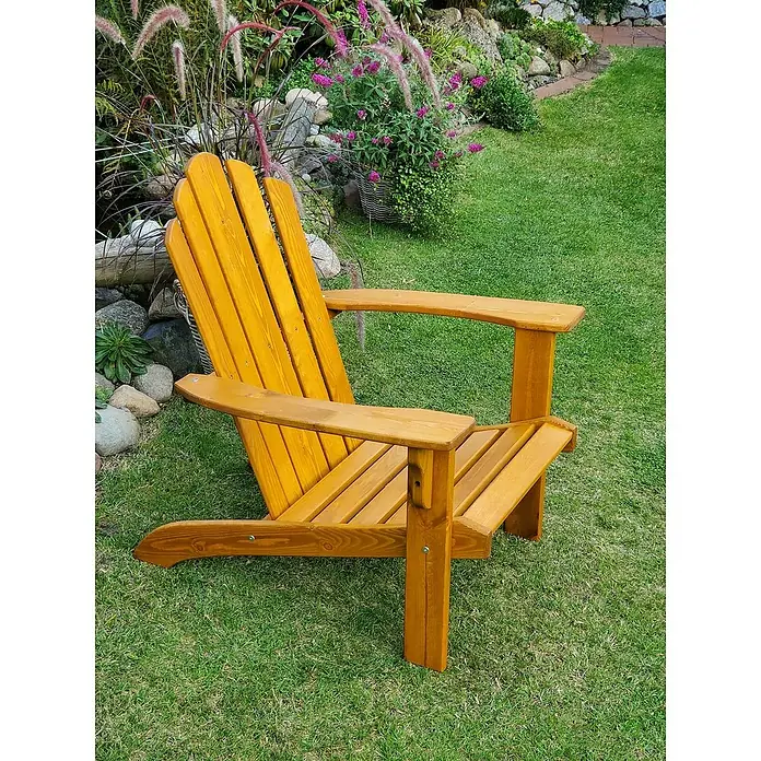 Promadino Gartenliege Adirondack - Farbe: Honigbraun - Ein goldener Adirondack-Stuhl aus Holz steht auf einem Rasen vor einem Garten mit Blumen. Ideal zum Entspannen im Freien.