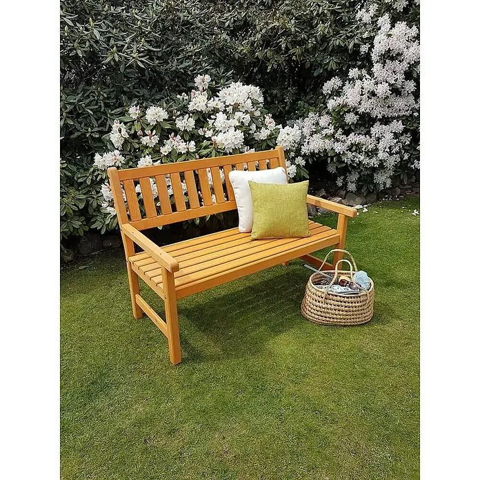 Promadino Gartenbank Johanna - B x T x H: 125 x 71 x 88,5 cm, Kiefer, Honigbraun - Eine Holzbank mit Kissen und einem Korb mit Strickzeug vor einem blühenden Busch. Die Gartenbank lädt zum Verweilen ein.