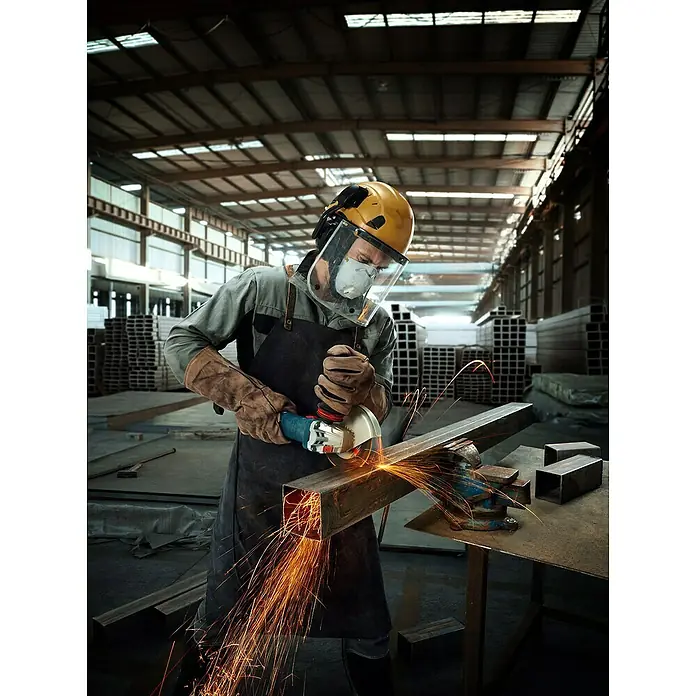 Bosch Professional Elektro-Winkelschleifer GWS 12-125 S - 1.200 W, Durchmesser Scheibe: 125 mm - Ein Arbeiter mit Schutzhelm und Schutzkleidung benutzt einen Winkelschleifer in einer Werkstatt. Funken sprühen während der Arbeit mit dem Winkelschleifer.