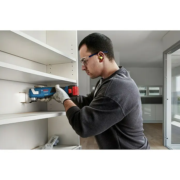 Bosch Professional AMPShare 18V Akku-Multifunktionswerkzeug GOP 18V-34 - 18 V, Ohne Akku - Ein Handwerker benutzt einen Bosch Professional GOP 18V-28 Akku-Multifunktionsschneider an einem weißen Regal, geschützt durch Brille und Gehörschutz.