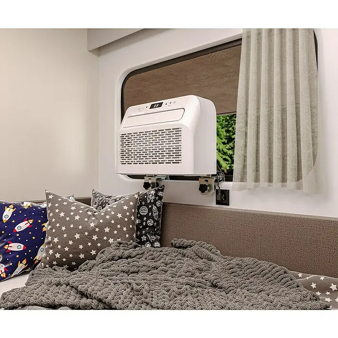 Voltomat Klimasplitgerät Camping - Kühlleistung: 500 W - 1.500 W, Raumgröße: 10 m² - 15 m² - Ein Fensterklimagerät ist an einem Fensterrahmen über einem Bett mit Kissen und einer Decke befestigt.