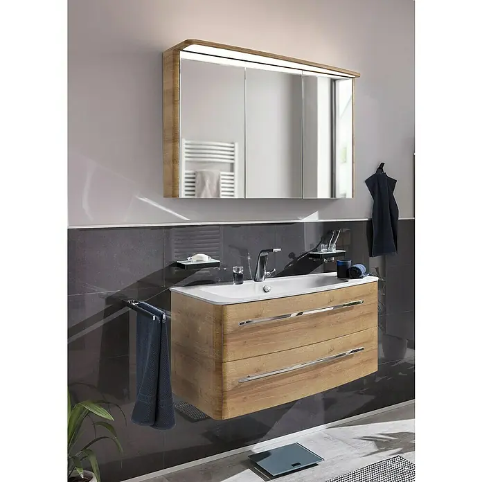 Lanzet Classico Badmöbel-Set CL10RWBC - 99,2 cm, Berg Eiche, Farbe Griff: Chrom, Rechts - Ein Badezimmer-Interieur mit einem Waschtischunterschrank aus Holz, einem weißen Aufsatzwaschbecken und einem Spiegelschrank mit integrierter Beleuchtung.
