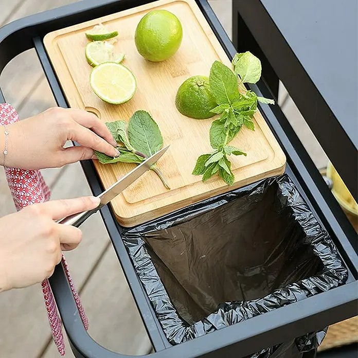 Mesa de barbacoa Reva 110 Premium para barbacoa - L x An x Al: 65 x 121 x 110 cm - Una tabla de cortar en un carrito de servicio con limas y menta, una persona corta menta sobre un bote de basura.