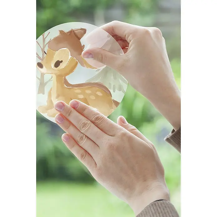 Komar Fenstersticker Disney Enchanted Forest Bubbles - 30 x 30 cm - Eine Person bringt einen Fensteraufkleber mit einem Waldtier-Motiv an, um eine winterliche und festliche Atmosphäre zu schaffen.