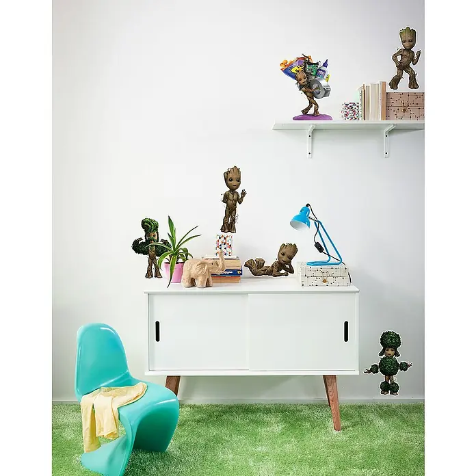 Komar Marvel Dekosticker I am Groot! - 70 x 50 cm - Eine verspielte Szene mit Groot-Figuren, die auf einem Sideboard und einem Regal platziert sind, ergänzt durch einen türkisfarbenen Stuhl und einen grünen Teppich.