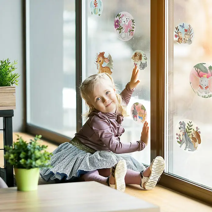 Komar Fenstersticker Disney Enchanted Forest Bubbles - 30 x 30 cm - Mädchen sitzt am Fenster mit Fensteraufklebern mit Tiermotiven. Das Sonnenlicht scheint durch das Fenster und beleuchtet die Szene.
