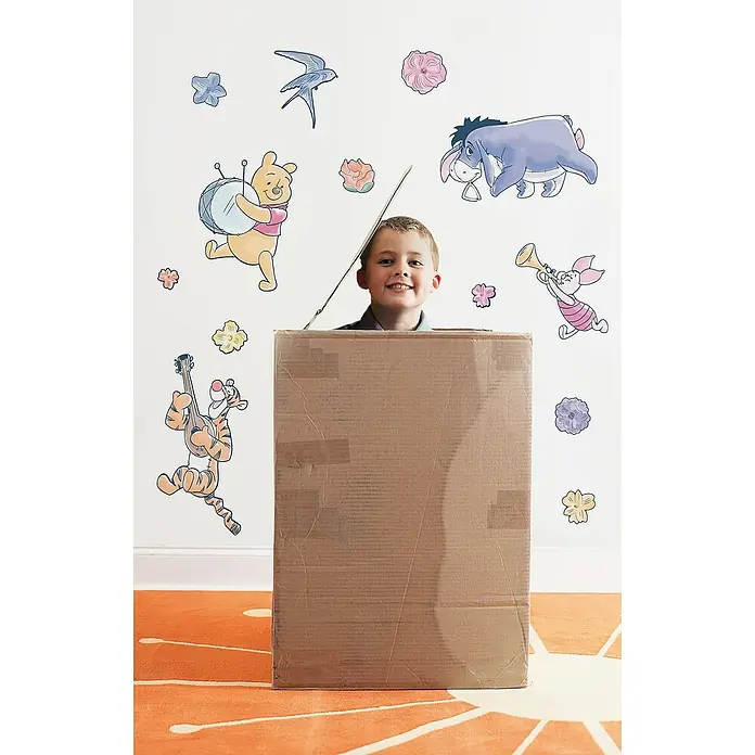 Komar Winnie Puuh Dekosticker Winnie the Pooh Rock-a-bye - 70 x 50 cm - Ein lächelnder Junge steht in einem großen Pappkarton vor einer Wand mit Winnie Puh-Motiven.