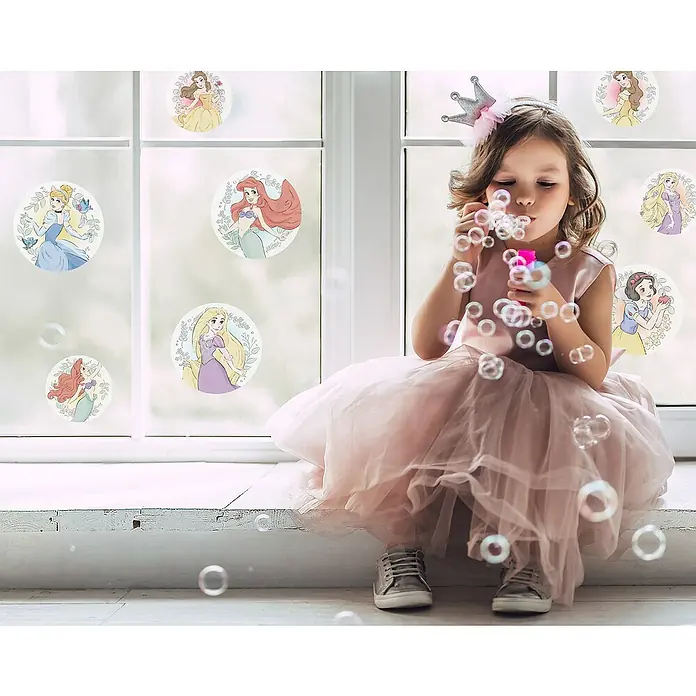 Komar Fenstersticker Disney Princess Kindness Bubbles - 30 x 30 cm - Ein Mädchen in einem rosa Kleid mit einer Krone bläst Seifenblasen vor einem Fenster mit Disney-Prinzessinnen-Aufklebern.
