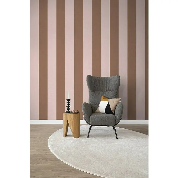 SCHÖNER WOHNEN-Kollektion Cosyliving Fototapete Streifen - B x H: 212 x 300 cm, Vlies, Braun-Rosa - Einladender grauer Sessel mit dekorativem Kissen auf einem weichen, runden Teppich, akzentuiert durch einen Holzhocker mit Kerzenständer vor gestreifter Wand.