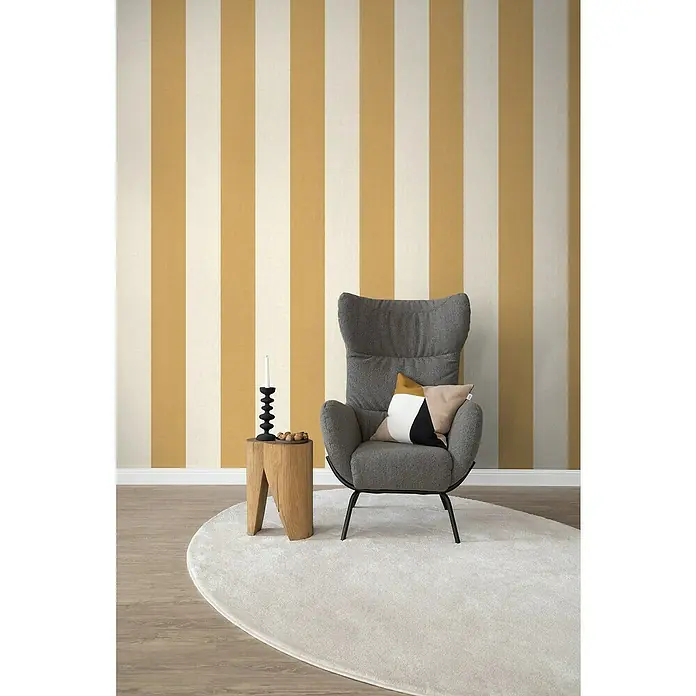 SCHÖNER WOHNEN-Kollektion Cosyliving Fototapete Streifen - B x H: 212 x 300 cm, Vlies, Gelb-Creme - Grauer Sessel mit Kissen auf einem cremefarbenen Teppich vor einer gestreiften Wand.