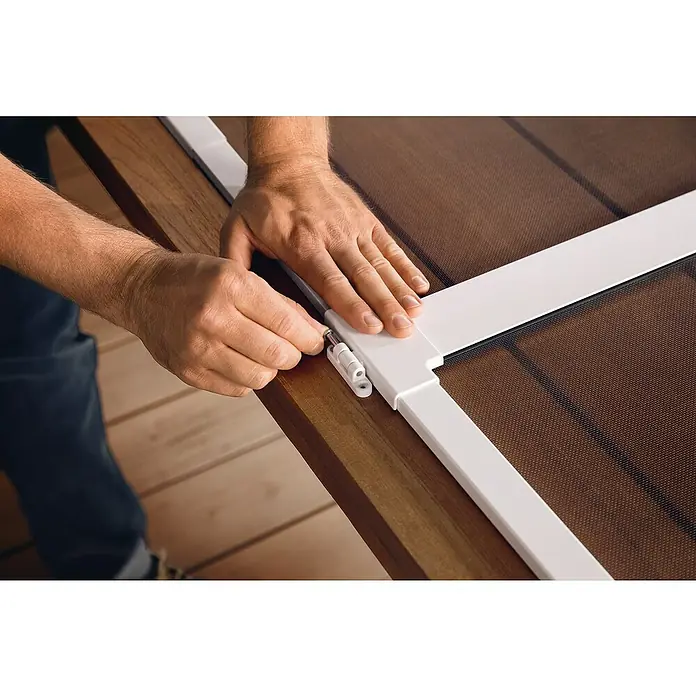 Tesa Puerta con protección contra insectos Falt - Antracita, Montaje de la mosquitera: Pinzas - Primer plano de una persona que fija una mosquitera blanca a un marco de madera, destacando el montaje de la mosquitera.