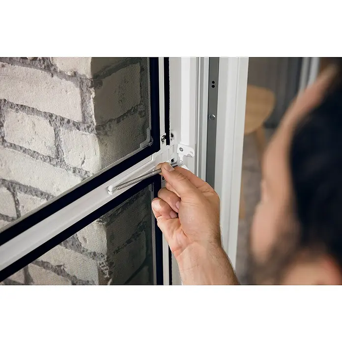 Tesa Insect Stop Puerta con protección contra insectos Falt - Blanco, Montaje de la mosquitera: Pinzas - Primer plano de una persona insertando un resorte en una puerta, presumiblemente una puerta mosquitera.