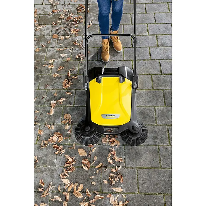 Kärcher Handkehrmaschine S 4 Twin - Flächenleistung: 2.400 m²/h, 20 l - Gelber Kärcher-Kehrmaschine im Einsatz auf einer gepflasterten Fläche voller Blätter, bedient von einer Person in Jeans und Stiefeln.
