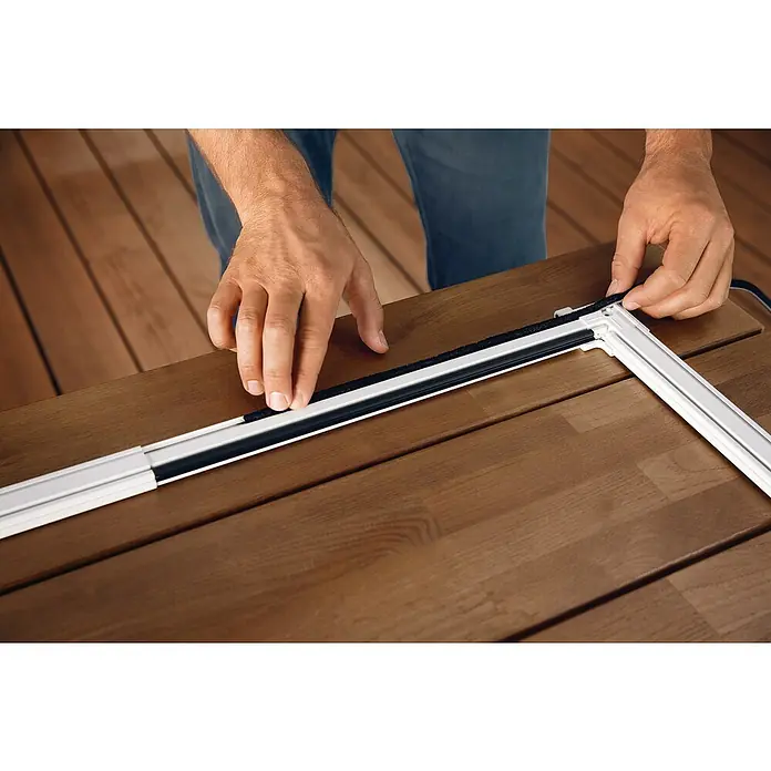 Tesa Puerta con protección contra insectos Falt - Antracita, Montaje de la mosquitera: Pinzas - Primer plano de una persona que fija un sello negro a un marco de ventana blanco, que está sobre una superficie de madera.