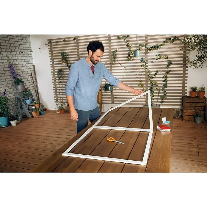 Tesa Insect Stop Puerta con protección contra insectos Falt - Blanco, Montaje de la mosquitera: Pinzas - Hombre construyendo un invernadero con perfiles de plástico blancos sobre una mesa de madera, con herramientas cerca.