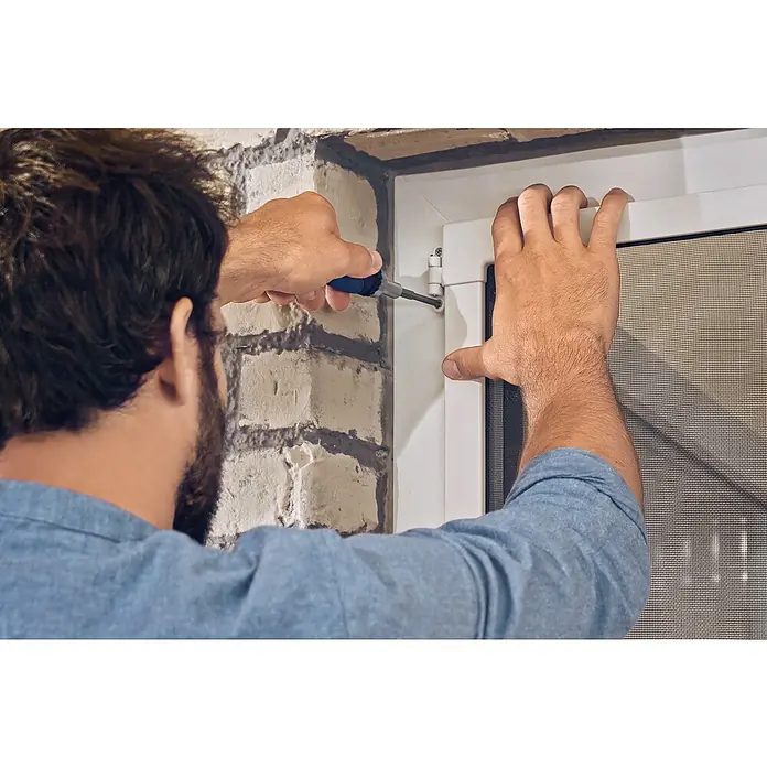 Tesa Puerta con protección contra insectos Falt - Antracita, Montaje de la mosquitera: Pinzas - Un hombre está fijando una ventana blanca con una mosquitera en una pared de ladrillos con un destornillador.