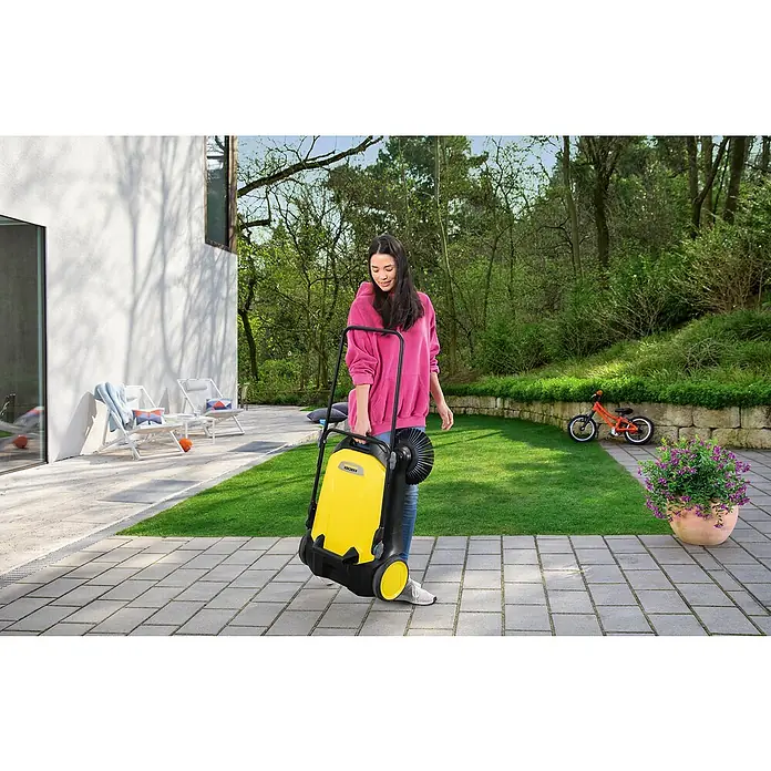 Kärcher Handkehrmaschine S 4 - Flächenleistung: 1.800 m²/h, 20 l - Eine Frau hievt eine gelbe Kärcher Kehrmaschine für den Außenbereich, um Terrassen und Wege sauber zu halten.