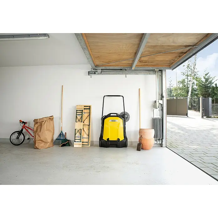 Kärcher Handkehrmaschine S 6 - Flächenleistung: 2.500 m²/h, 38 l - Eine gelbe Kehrmaschine steht in einer Garage neben Gartenwerkzeugen, einem Fahrrad und einem zusammenklappbaren Tisch.