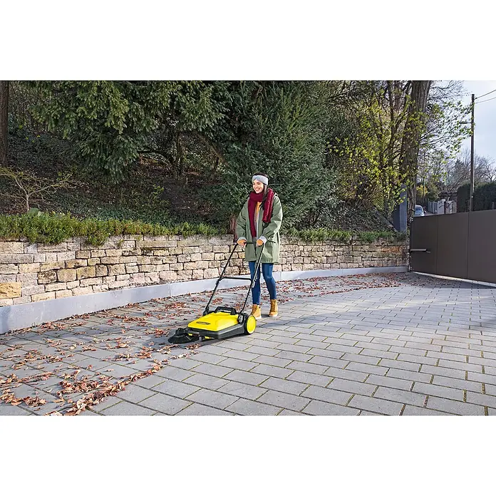 Kärcher Handkehrmaschine S 4 - Flächenleistung: 1.800 m²/h, 20 l - Frau kehrt mit der gelben Kärcher-Kehrmaschine Blätter auf einem gepflasterten Weg. Das Gerät ist ideal für Haus und Garten, um Flächen sauber zu halten.