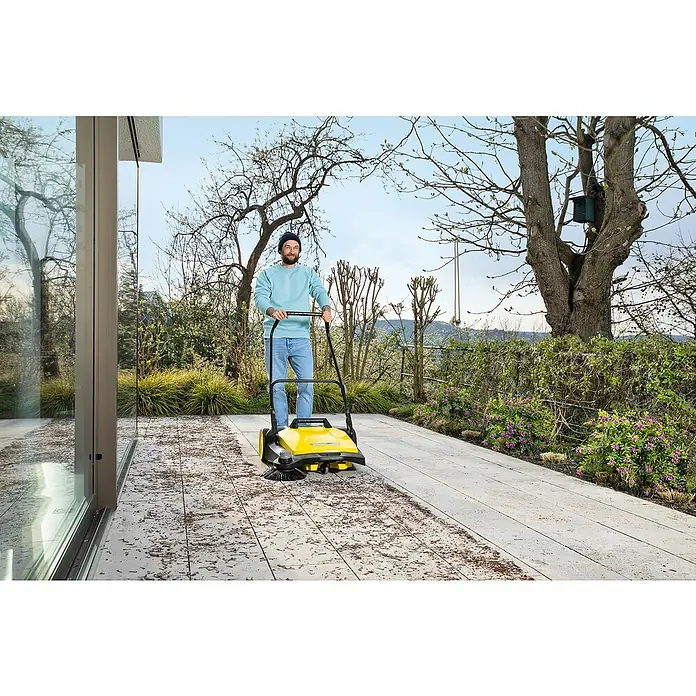 Kärcher Handkehrmaschine S 6 - Flächenleistung: 2.500 m²/h, 38 l - Ein Mann benutzt eine gelbe Kehrmaschine von Kärcher, um Laub von einer Terrasse zu entfernen. Die Kehrmaschine säubert effizient den Außenbereich.