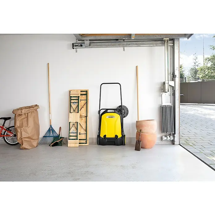 Kärcher Handkehrmaschine S 4 - Flächenleistung: 1.800 m²/h, 20 l - Das Bild zeigt eine Kärcher Kehrmaschine in einer Garage, neben anderen Gartengeräten wie einem Rechen und einem Besen.