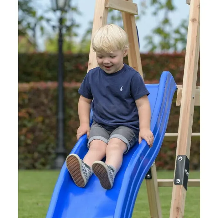 Swing King Doppelschaukel Ava - L x B x H: 73 x 245 x 170 cm, Holz, Blau - Ein lächelnder Junge rutscht eine blaue Kinderrutsche auf einem hölzernen Spielplatz herunter, wobei seine Füße im Vordergrund sind.