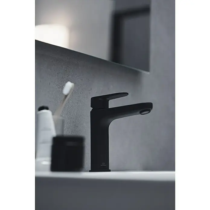 Ideal Standard Cerafine O Waschtischarmatur H120 - Schwarz, Matt, Mit Push-Open Ablaufgarnitur - Elegante Nahaufnahme eines schwarzen Waschtischhahns, Teil eines modernen Badezimmer-Designs, mit Zahnbürste und Pflegeprodukten im Hintergrund.