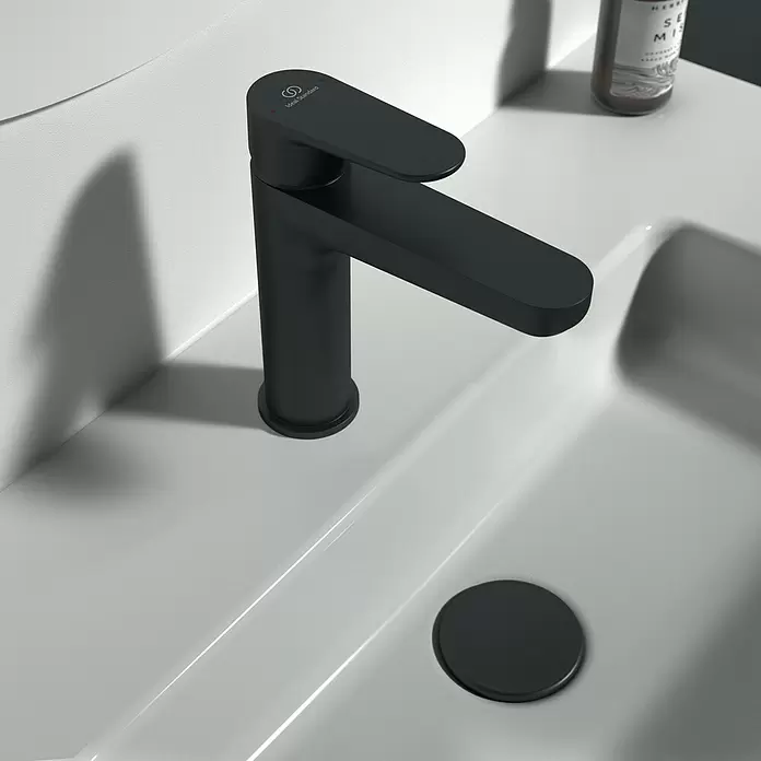 Ideal Standard Cerafine O Grifo de lavabo - Negro, Mate, Con juego de desagüe «Push-Open» - Grifo de baño negro mate con logo Ideal Standard, instalado en un lavabo blanco con tapa de desagüe negra a juego.
