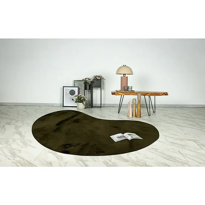 Fellteppich Happy - Basil, 230 x 160 cm, Oval, 100 % Polyester (Flor) - Ein stilvolles, unregelmäßig geformter olivgrüner Teppich liegt auf einem Marmorboden mit Möbeln und Deko im Hintergrund.